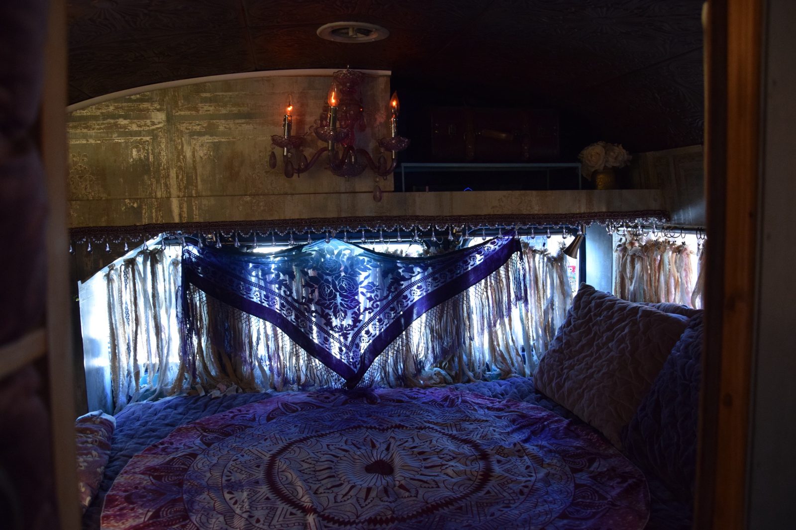 The bedroom at the rear of the bus: a mandala throw on the bed, ribbon-lace curtains, and a small crystal chandelier sconce