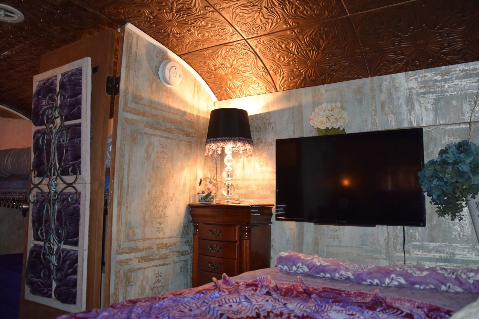 Bedroom corner with embossed copper ceiling tiles, a crystal lamp on the dresser, and a wall-mounted TV