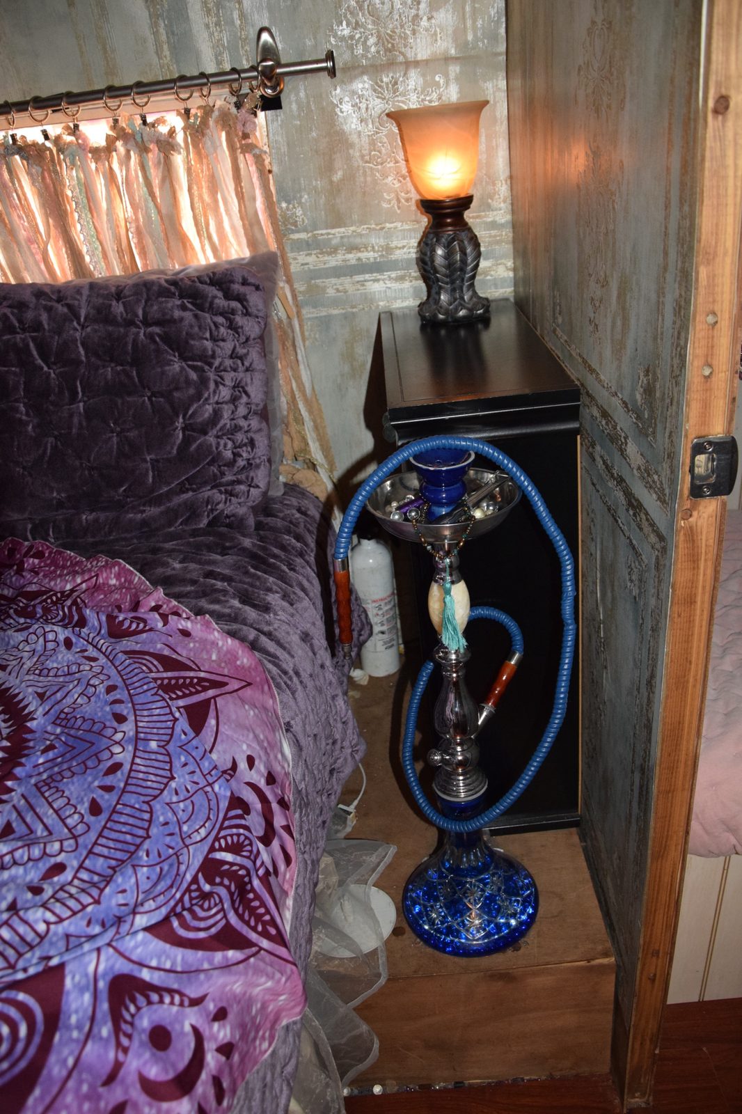 A blue glass hookah standing beside the bed's velvet quilt and lace curtains