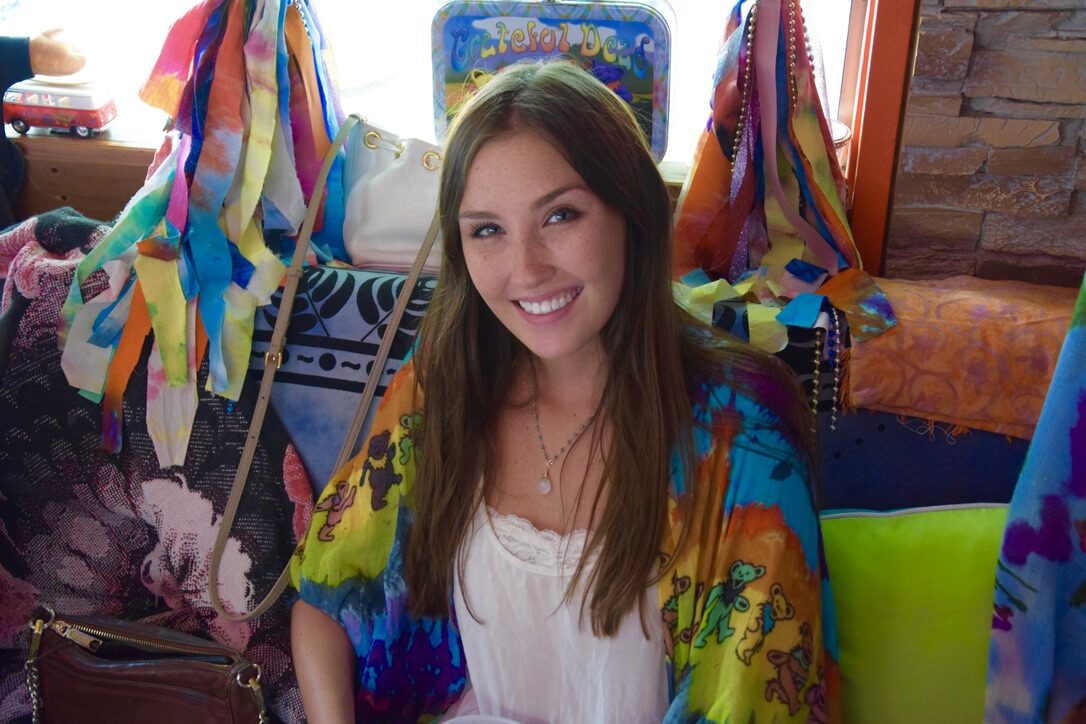 A young woman in a Grateful Dead dancing bears tie-dye jacket, seated inside SUGAREE