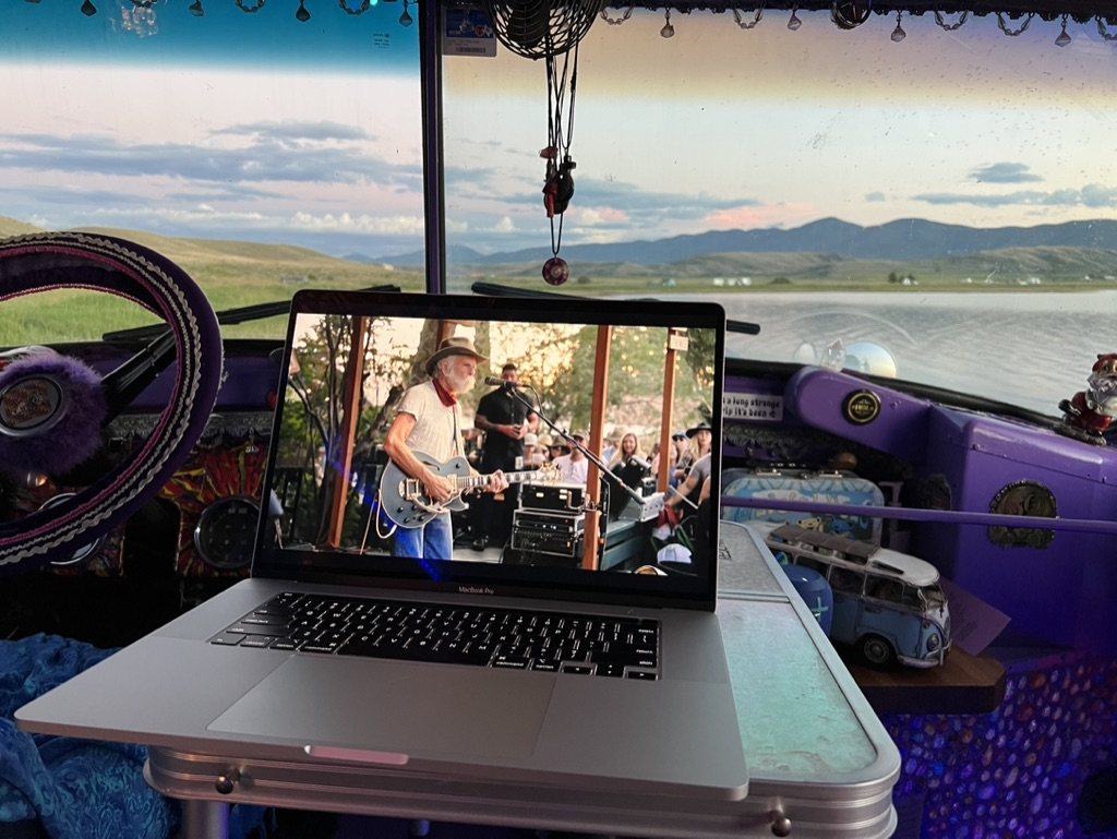 A laptop playing a show on the bus dash with a lake and mountains through the windshield