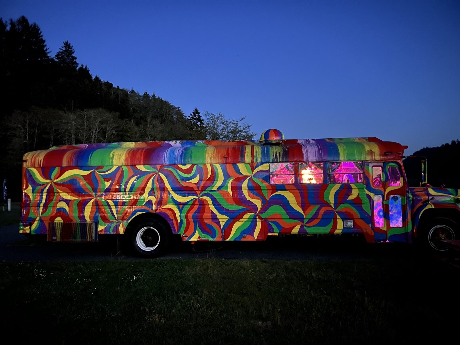 SUGAREE at dusk with pinwheel paint panels and glowing purple interior lights, parked in front of dark tree-covered hills