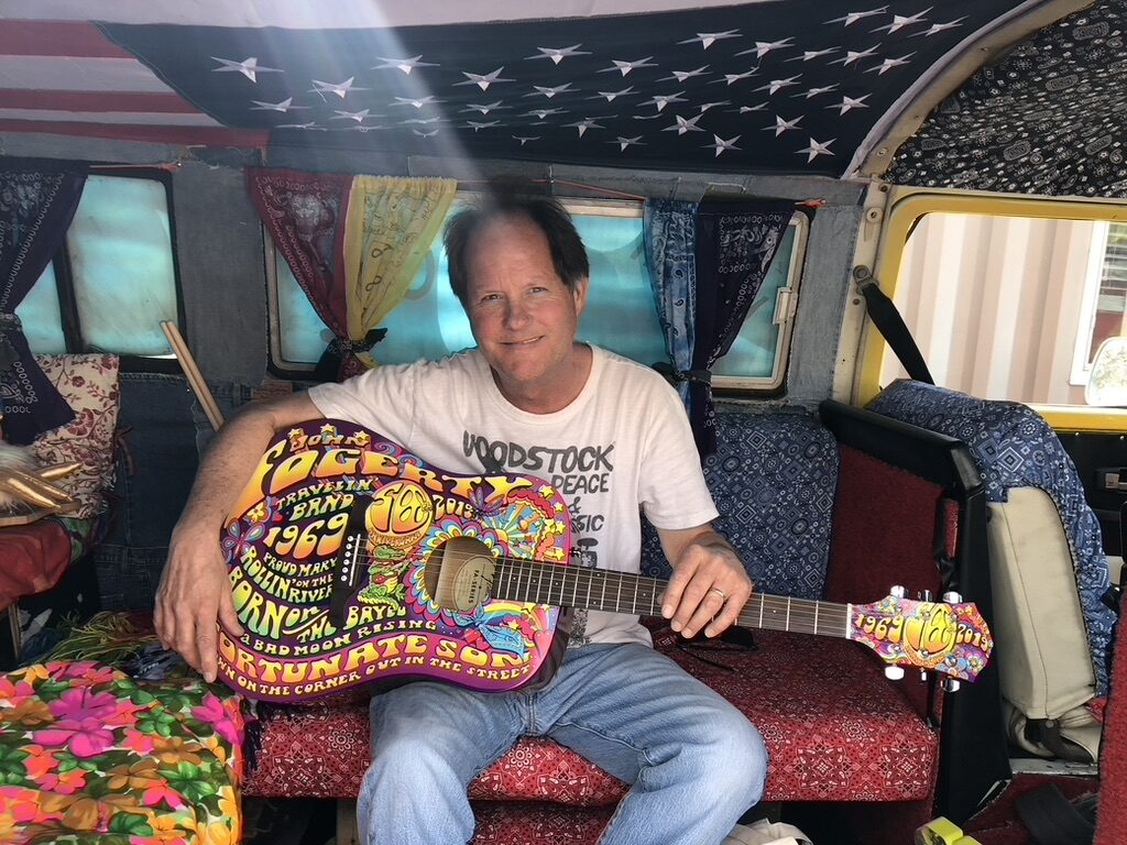 A man holding a custom John Fogerty tour guitar while sitting inside SUGAREE