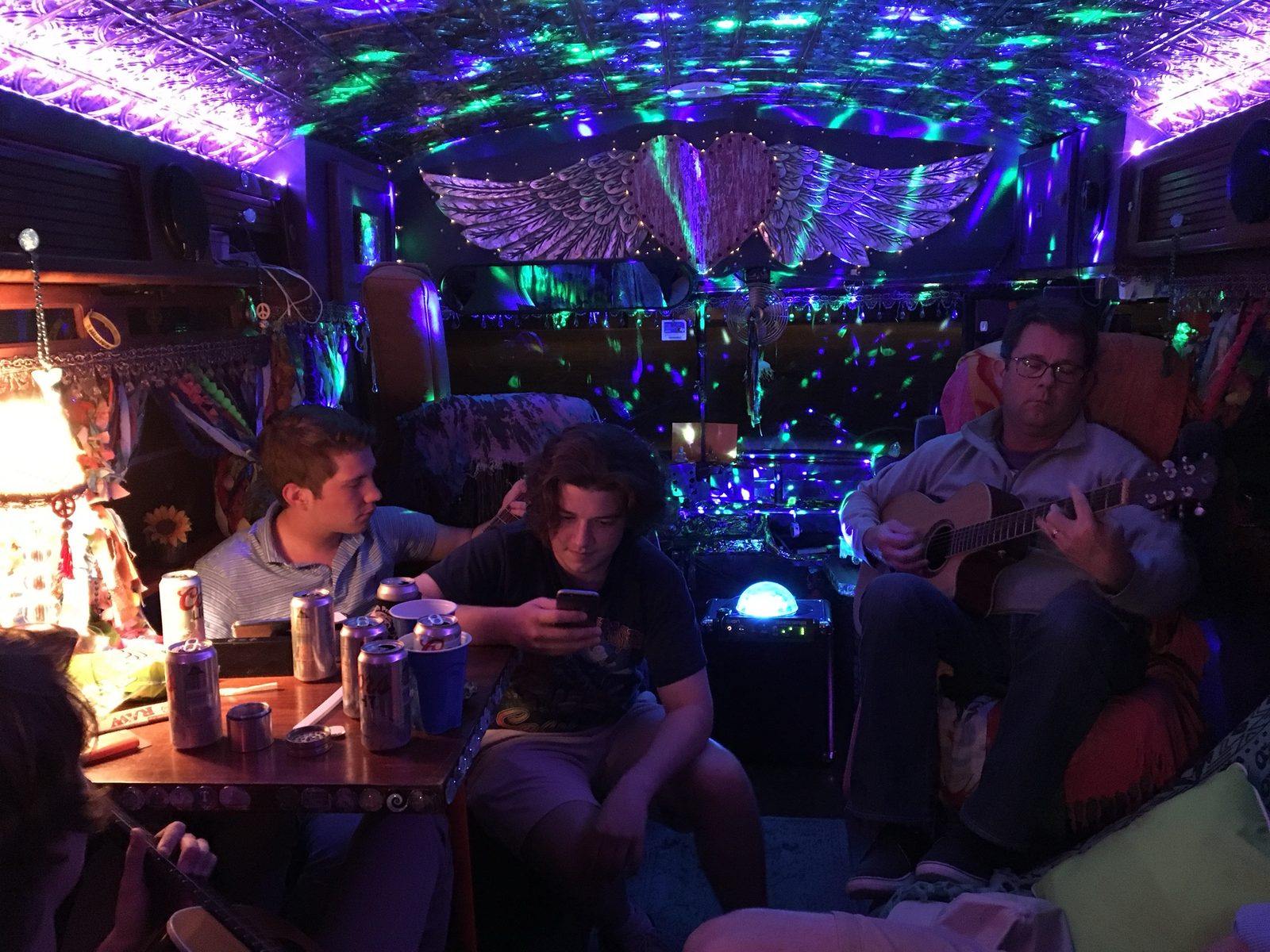 Friends gathered around the table at night while one plays acoustic guitar under green and purple party lights