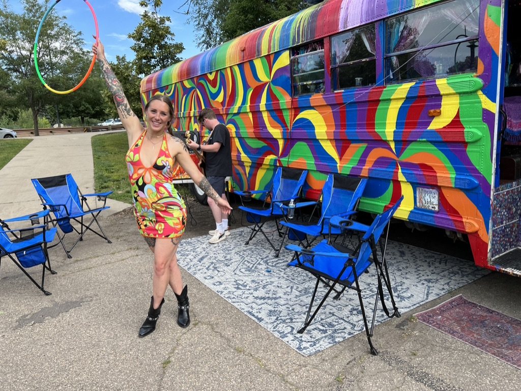 A woman holding a rainbow hula hoop beside SUGAREE with camp chairs set out