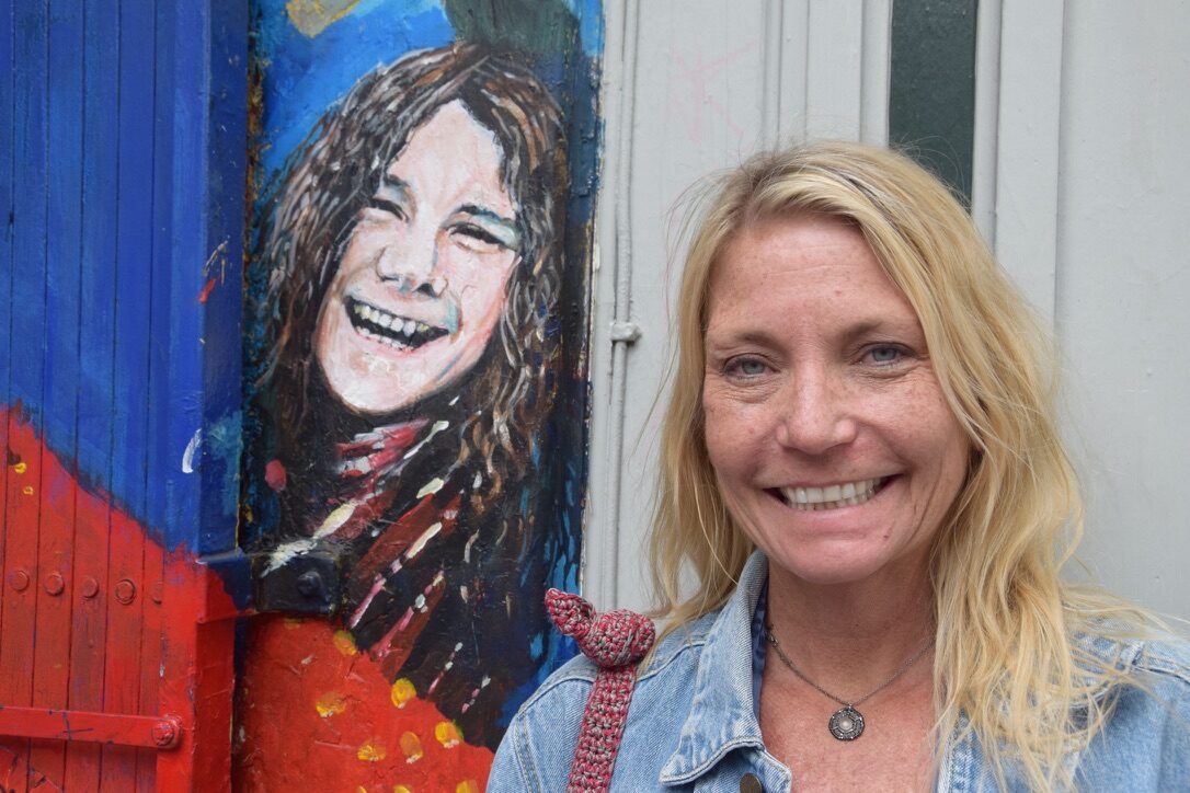 A woman standing beside a painted mural of Janis Joplin in the Haight-Ashbury district