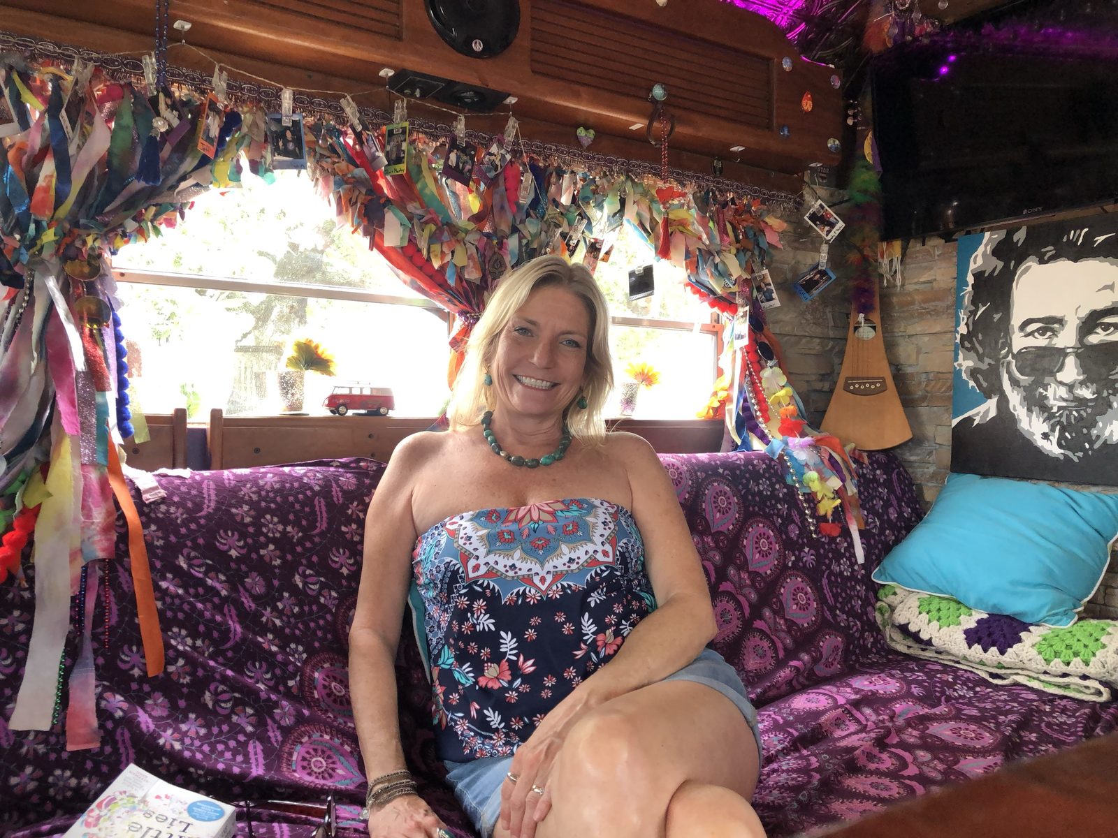 Kelly smiling on the purple paisley couch beneath ribbon curtains, a Jerry Garcia portrait on the wall beside her