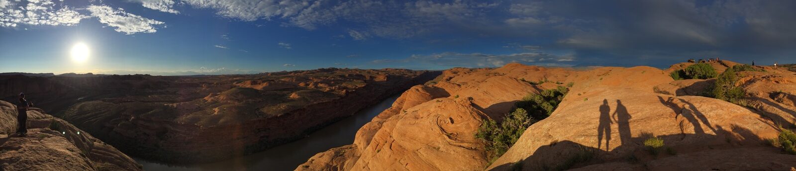 Panoramic view of a red rock canyon and river near Moab at sunset, shadows of two people cast on the rock