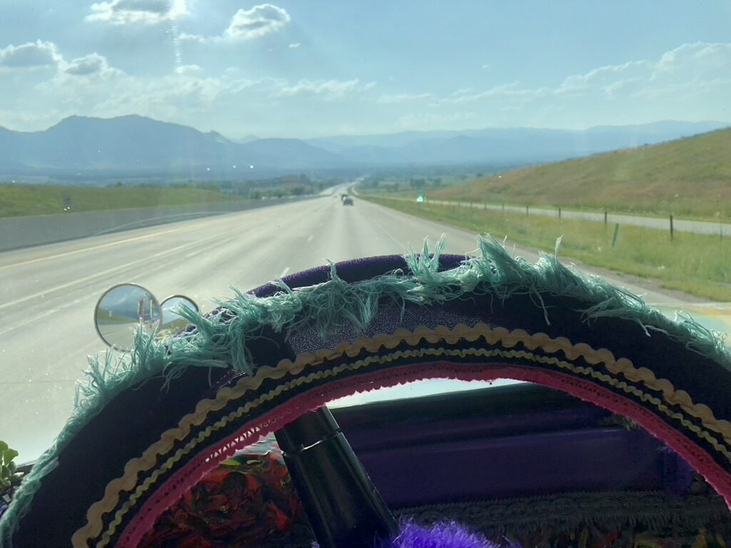 View through SUGAREE's windshield of a mountain highway, tasseled steering wheel cover in frame