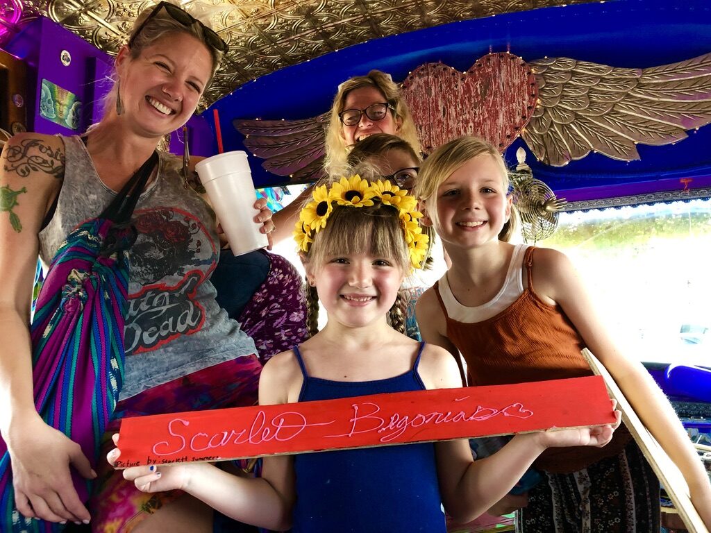 A woman and three girls inside SUGAREE holding a hand-painted 'Scarlet Begonias' sign, one wearing a sunflower crown