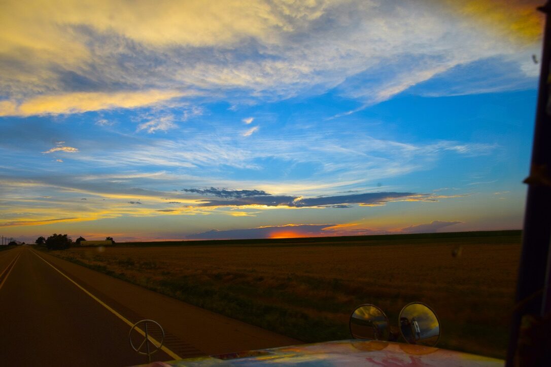 A wide sunset sky over open fields, seen from SUGAREE's cab with the peace-sign mirrors in view