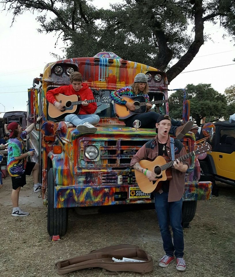 Three young men playing acoustic guitars on and around the hood of SUGAREE
