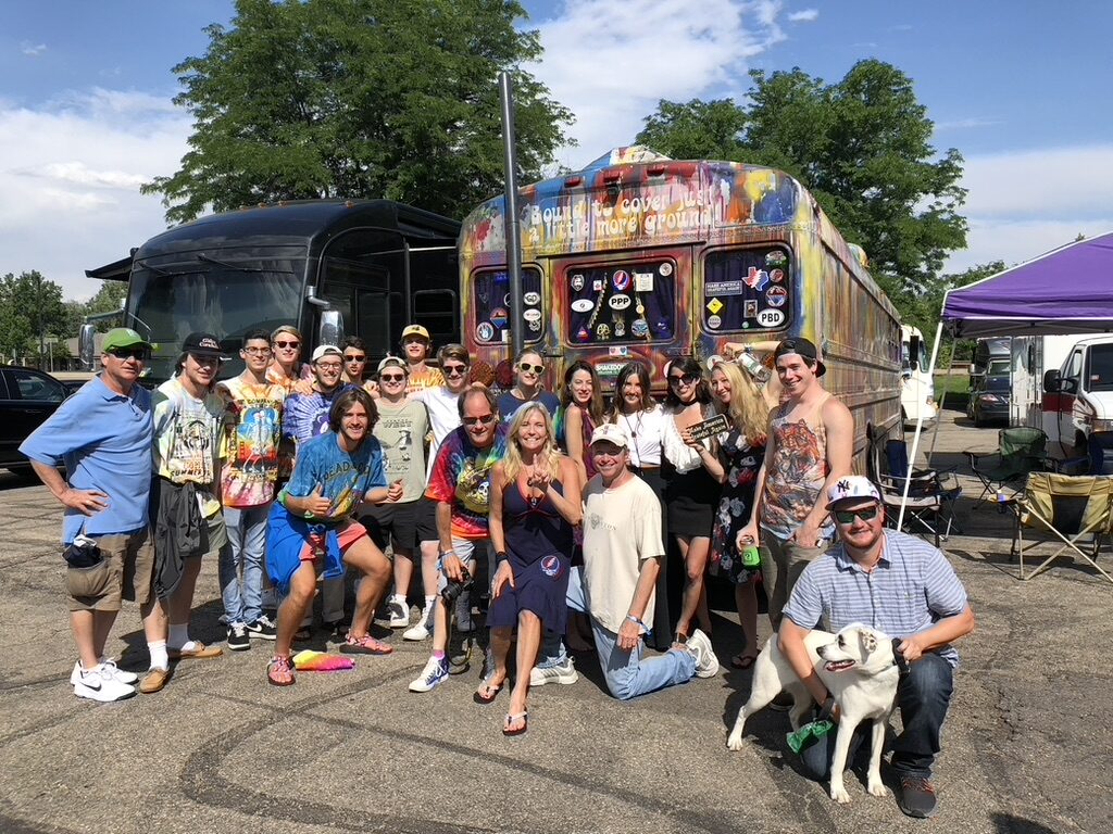 A large group posing in front of SUGAREE and another tour bus painted with 'Bound to cover just a little more ground,' a dog in front