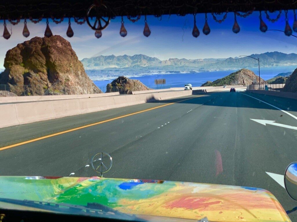 View through SUGAREE's windshield, peace sign hood ornament, driving along a coastal mountain highway